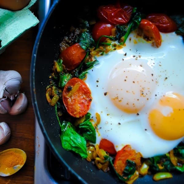 A pan of fried eggs with wilted spinach and blistered cherry tomatoes, next to eggs, onion and garlic.