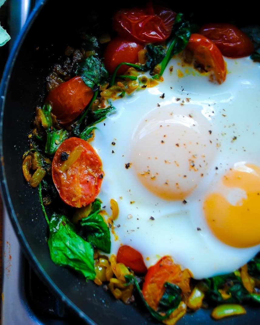 A pan of fried eggs with wilted spinach and blistered cherry tomatoes, next to eggs, onion and garlic.