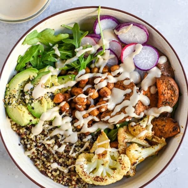 A grain bowl with roasted cauliflower, chickpeas, avocado, radish and greens under a tahini drizzle.