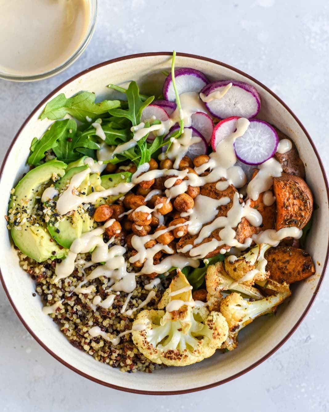 A grain bowl with roasted cauliflower, chickpeas, avocado, radish and greens under a tahini drizzle.