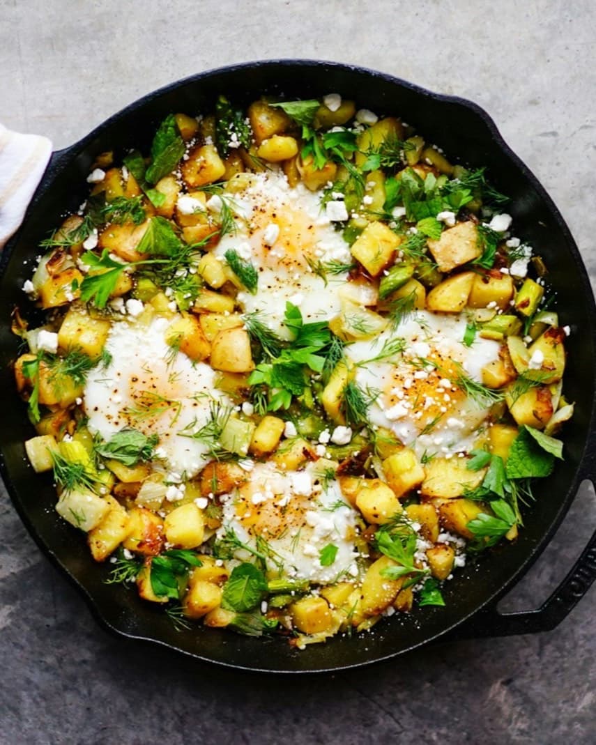 A cast-iron skillet of eggs baked into greens, crisp potatoes and crumbled feta, seen from above.