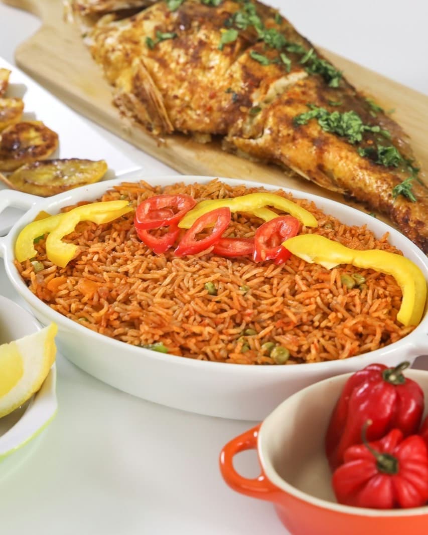 A dish of jollof rice with grilled fish, skewers, lemon wedges and fresh peppers.