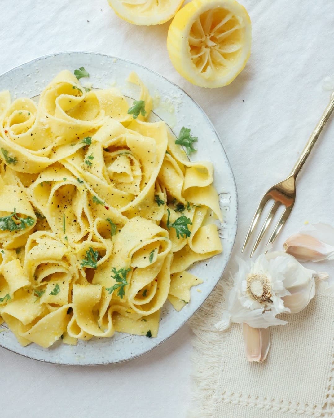 A plate of buttery lemon pappardelle with a fork, squeezed lemon halves and garlic on a white cloth.