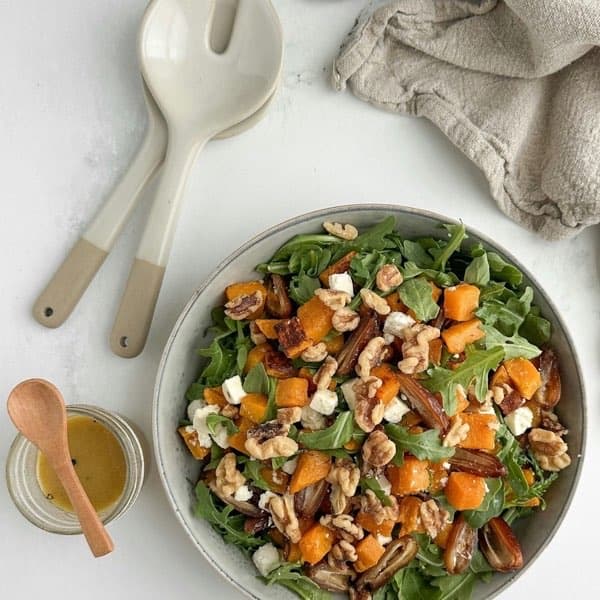 A bowl of arugula with roasted butternut squash, walnuts and feta, serving spoons and dressing beside it.
