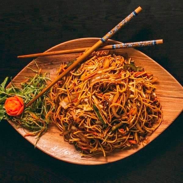 Stir-fried noodles with vegetables on a wooden platter with chopsticks.