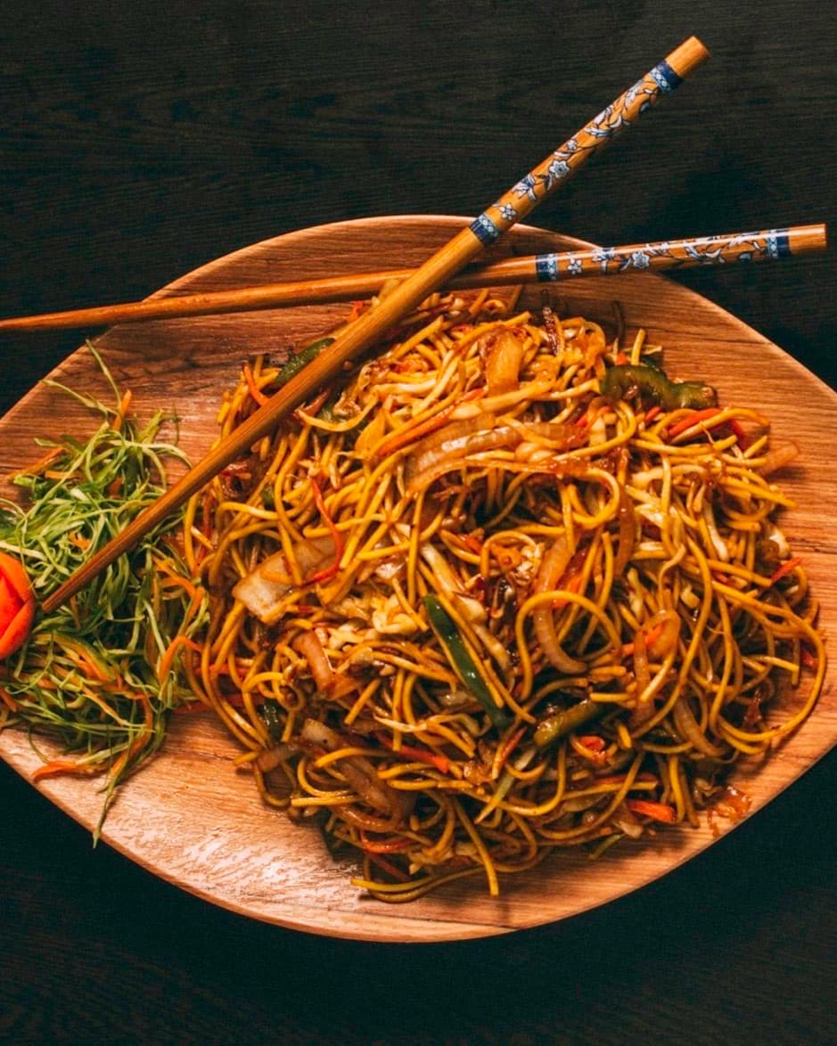 Stir-fried noodles with vegetables on a wooden platter with chopsticks.